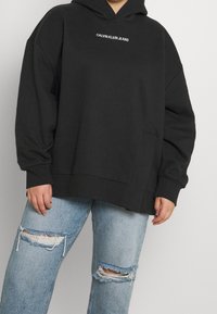 Felpa nera over size con tasca frontale e logo bianco "CALVIN KLEIN JEANS". Abbinata a jeans strappati blu chiaro.