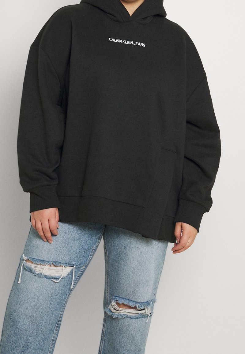 Felpa nera over size con tasca frontale e logo bianco "CALVIN KLEIN JEANS". Abbinata a jeans strappati blu chiaro.