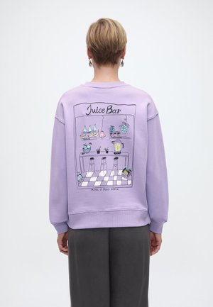 Personne aux cheveux blonds courts portant un sweat-shirt lavande avec une illustration colorée d'un bar à jus au dos, comprenant des fruits et des tabourets.