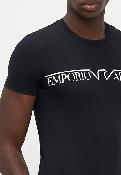 Melna kokvilnas t-kreklu ar īsām piedurknēm, uz kura krūšu daļas ir balts "EMPORIO ARMANI" logo. Tuvināta piegriezuma dizains ar gludu tekstūru.