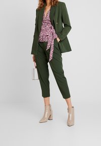 Olivgrön skräddarsydd kavaj över en blommig peplumtopp med rosa och vita mönster, ihop med gröna smalrandiga byxor och beige ankelskor.