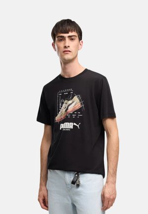 Giovane uomo con una t-shirt nera Puma con grafica di sneaker e jeans azzurri, in piedi davanti a uno sfondo bianco neutro.