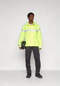 Ryški geltona neperšlampama striukė su atšvaitinėmis juostelėmis, juodos kelnės ir juodi batai; modelis laiko dviračio šalmą.