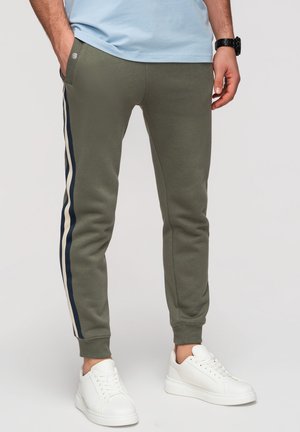 Homme portant un jogging vert olive avec des bandes latérales bleu marine et blanches, une chemise bleu clair, des baskets blanches et une montre-bracelet noire, mains détendues.