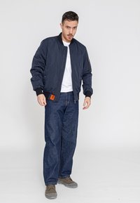Maritieme bomberjack met een ritszak, geribde manchetten en een oranje label. Gecombineerd met blauwe denimspijkerbroek en grijze sneakers op een neutrale achtergrond.