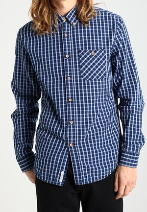 Man draagt een overhemd met blauwe en witte ruitjes, een borstzak en houten knopen, staat met ontspannen handen langs zijn zij.