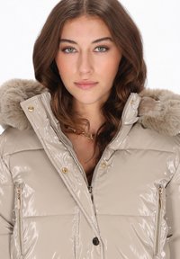 Geacă matlasată lucioasă bej cu glugă captusită cu blană artificială, buzunare cu fermoar și accesorii în nuanță aurie. Are panouri cusute și textură lucioasă.