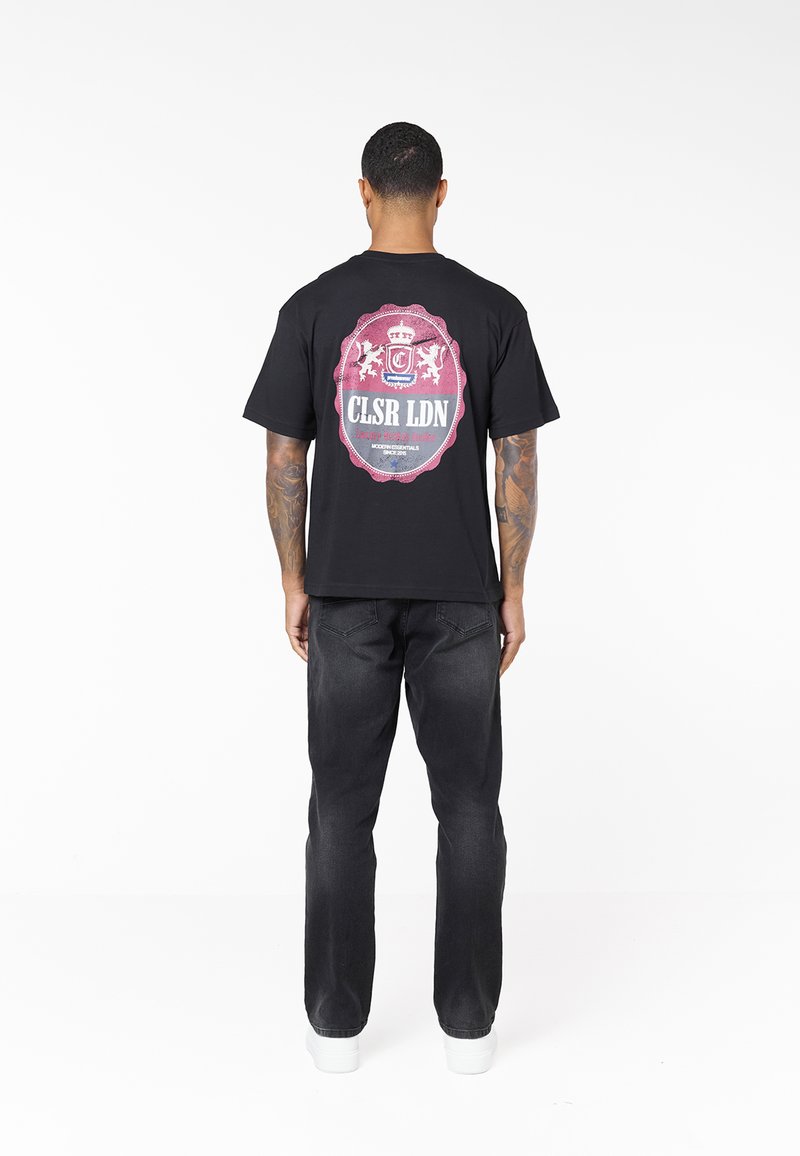 Muž s tetováním stojí zády, má na sobě černé tričko s potiskem, tmavé džíny a bílé tenisky na bílém pozadí.