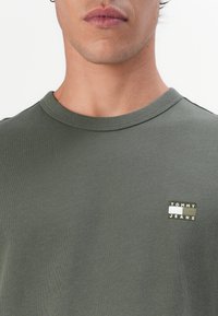 Tricou din bumbac de culoare verde măsliniu cu guler rotund. Prezintă un mic patch brodat "TOMMY JEANS" în alb și verde închis. Textură netedă.