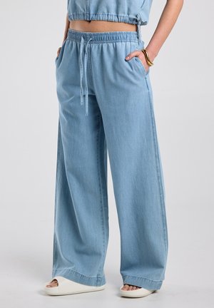 Vrouw in lichtblauwe wijde broek met trekkoord, witte slippers en een gouden armband, staand met handen in de zakken.