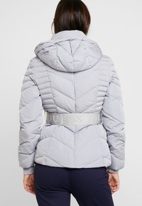 Femme vue de dos portant une veste matelassée gris clair avec une capuche et une large ceinture avec l'inscription "GUESS", associée à un pantalon foncé.