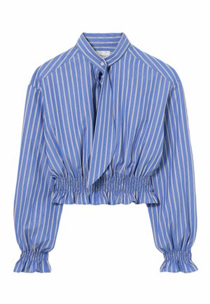 Donna in piedi che indossa una camicetta a righe blu e bianco con vita e polsini arricciati, abbinata a pantaloni larghi marroni a vita alta e scarpe marroni.