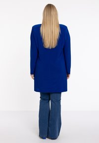 Frau mit langen blonden Haaren, die einen königsblauen Mantel und flared blaue Jeans trägt, steht mit dem Rücken zu einem weißen Hintergrund.