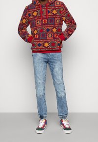 Červená vzorovaná flísová mikina s kapucí a šňůrkou, spárovaná s světle modrými roztrhanými džíny a barevnými teniskami s bílými tkaničkami.