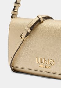 Sac bandoulière doré métallique en simili cuir texturé, avec une sangle réglable et des accessoires dorés. Il présente un logo embossé à l'avant.