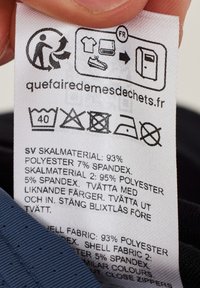 Pflegeetikett mit Waschsymbole, weißer Stoff, Text in Schwedisch und Französisch, der die Materialzusammensetzung und Pflegehinweise angibt.