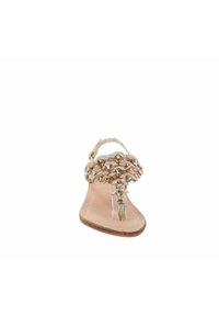 Sandalo piatto beige con cinturino intrecciato in ecopelle decorato con ornamenti color oro e un cinturino alla caviglia trasparente per una vestibilità sicura.