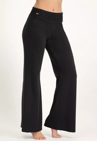 Kvinna iklädd svarta vida byxor med ett högt, elastiskt midjeband, som står barfota mot en enkel ljus bakgrund.