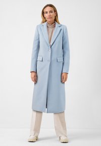 Lichtblauwe wollen jas met een uitgesneden kraag, twee voorzakken en drie knopen. Gecombineerd met beige wijde broek en chunky witte schoenen.