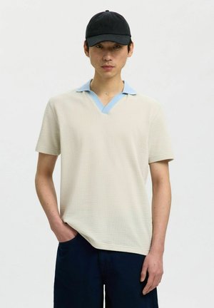 Giovane uomo che indossa una camicia a maniche corte color crema con trama, colletto azzurro chiaro e un berretto da baseball nero, in piedi con una mano in tasca.