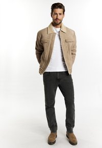 Beige Wildlederjacke mit Schafslederkragen, Druckknöpfen, zwei Brusttaschen, getragen über einem weißen Hemd und kombiniert mit schwarzen Jeans und braunen Stiefeln.