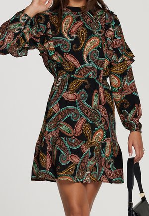Femme portant une robe noire avec des motifs cachemire colorés et des détails à volants, tenant un sac à main noir dans sa main droite.