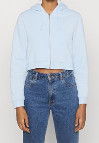 Ljust blå cropped hoodie med dragkedja framtill, snörning i huvan och ribbade muddarna, kombinerad med högmidjade blå jeans.