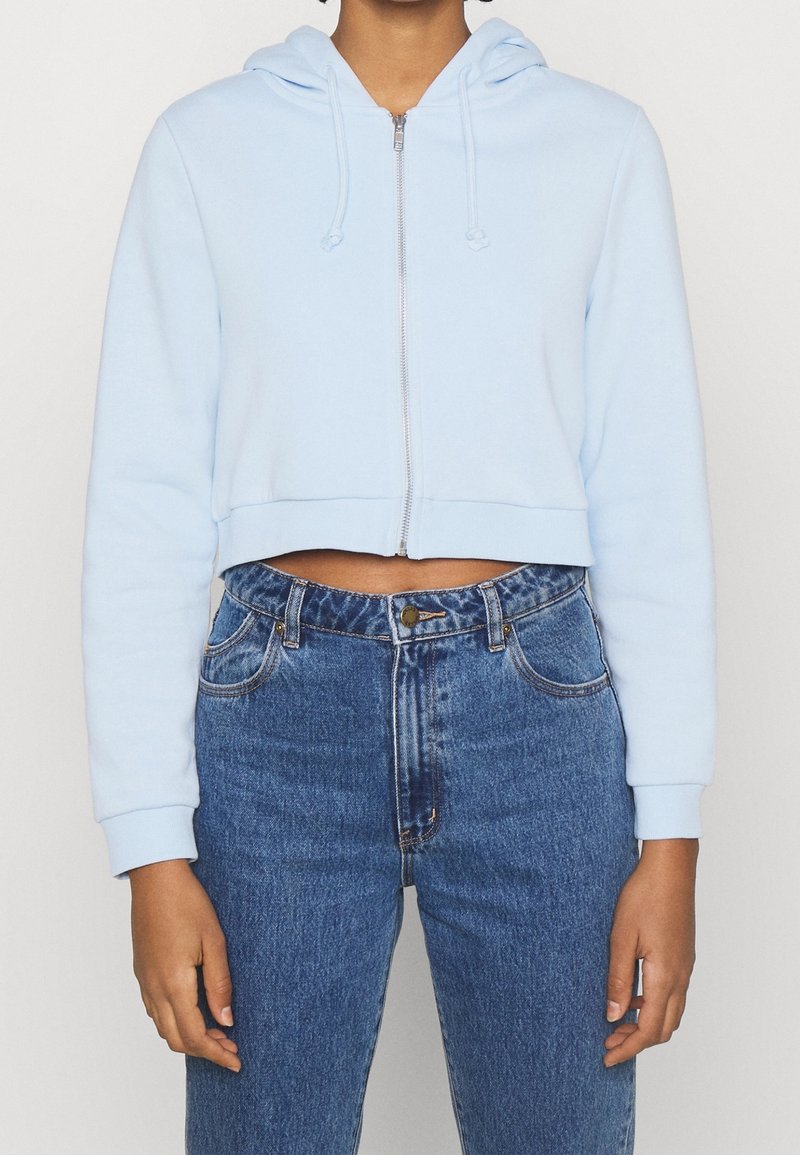 Ljust blå cropped hoodie med dragkedja framtill, snörning i huvan och ribbade muddarna, kombinerad med högmidjade blå jeans.