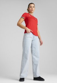 Rdeča športna majica z črnimi in belimi detajli, ki ima teksturiran vzorec, v kombinaciji z svetlo modrimi širokimi hlačami in črnimi športnimi copati.