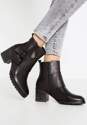 Bottes noires en cuir jusqu'aux chevilles avec talons carrés, fermetures éclair latérales et boucles décoratives, portées avec un jean gris clair sur un fond blanc.