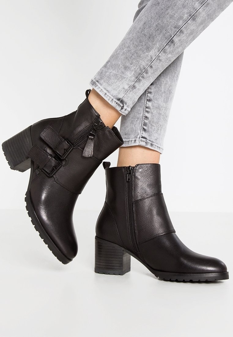 Bottes noires en cuir jusqu'aux chevilles avec talons carrés, fermetures éclair latérales et boucles décoratives, portées avec un jean gris clair sur un fond blanc.
