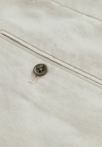Beige kangas, jossa on hieno rakenne, sisältää yhden tumman napin ja pienen ommelvarauksen. Materiaali on hieman rypistynyt.
