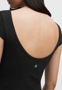 Haut noir ajusté avec un décolleté plongeant à l'arrière et des manches courtes, avec un petit logo dans le dos. Tissu lisse et extensible avec un léger éclat.