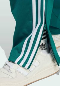 Donji dijelovi nogu u tirkiznim trenirama s bijelim prugama i bočnim patentnim zatvaračem, bijelim Adidas tenisicama i bijelim čarapama s crnim prugama i logom.
