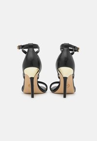 Para czarnych sandałów na wysokim obcasie z cienkimi paskami wokół kostki i złotym akcentem z tyłu obcasów, pokazana z tyłu na białym tle.