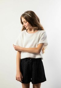 Biały, teksturowany sweter z krótkim rękawem w połączeniu z czarnymi szortami z subtelnym kropkowanym wzorem. Modelka stoi, ma jasnobrązowe włosy.