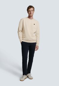Beige katoenen sweatshirt met ronde hals, geribde manchetten en tailleband. Draagt bij donkerblauwe broek en lichtgekleurde sneakers. Circulaire logo-detail op de borst.