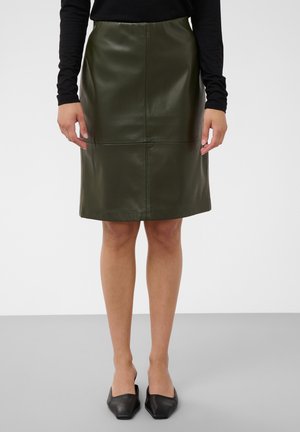 Femme portant une jupe en cuir vert olive foncé longueur genou, un haut noir à manches longues et des chaussures plates noires à bout pointu, debout sur un sol gris.