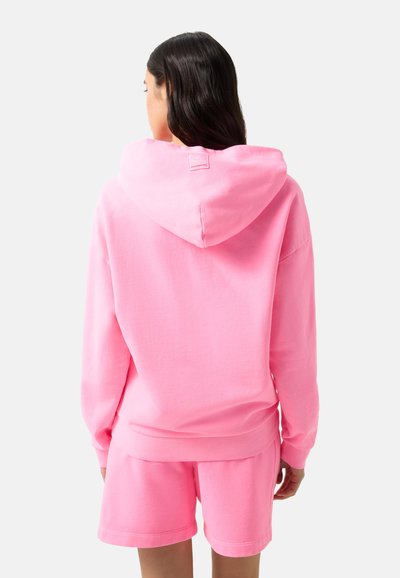 Femme aux longs cheveux foncés portant un sweat à capuche rose vif et un short assorti, vue de dos sur un fond blanc.