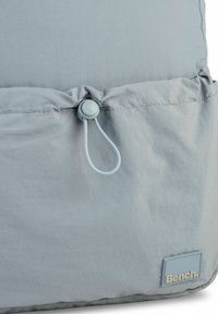 Helles graues Stoffbeutel mit einem Zugverschluss, versammeltem Nahtdetaill und einem kleinen blauen Etikett mit der Aufschrift "Bench". Das strukturierte Material ist sichtbar.