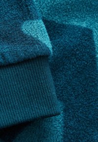 Strukturierter türkisfarbener Stoff mit einer weichen, flauschigen Oberfläche und einem gerippten Strickbund. Enthält unterschiedliche Blautöne in einem geschichteten Muster.