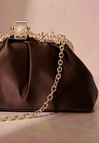 Dark brown satin evening bag with golden floral clasp and chain strap on a light pink surface.