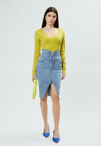 Top verde a maniche lunghe con scollatura a V, gonna di jeans blu a vita alta con spacco frontale, tacchi blu a punta e borsa verde.