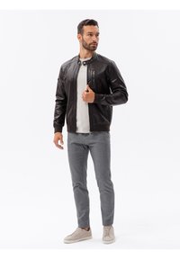 Giacca bomber in pelle marrone con colletto rialzato, tasca sul petto con zip e polsini a costine, abbinata a pantaloni grigio chiaro e sneakers beige.