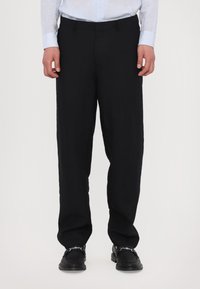 Schwarze Leinenhosen mit lässiger Passform, flacher Vorderseite und Gürtelschlaufen, kombiniert mit schwarzen Schuhen mit dekorativen Hardware-Details.