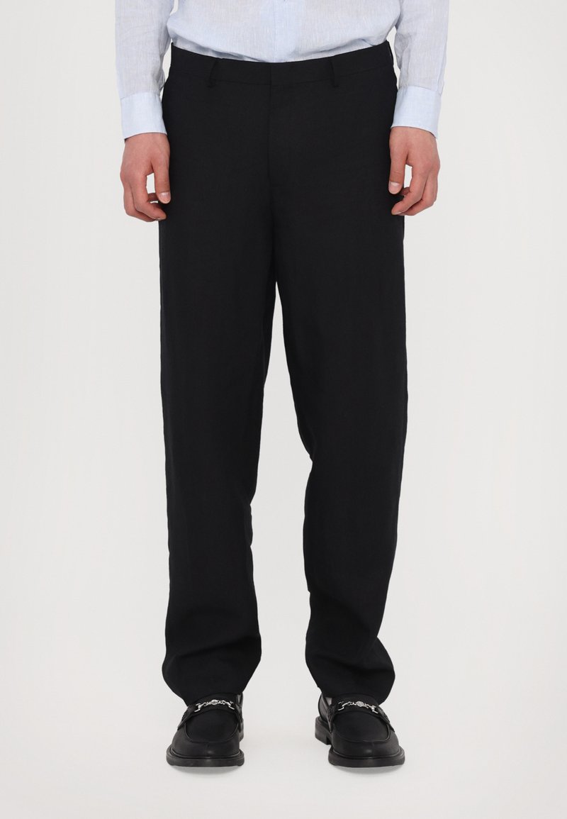 Schwarze Leinenhosen mit lässiger Passform, flacher Vorderseite und Gürtelschlaufen, kombiniert mit schwarzen Schuhen mit dekorativen Hardware-Details.