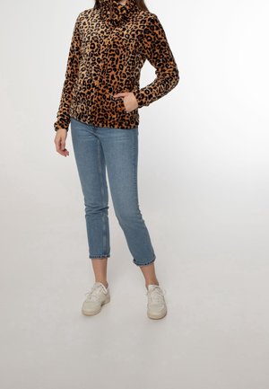 Woman standing wearing a brown leopard-print fleece jacket, light blue cropped jeans, and white sneakers, hand in jacket pocket.