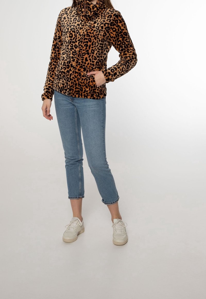 Femme debout portant une veste polaire marron à imprimé léopard, un jean court bleu clair et des baskets blanches, main dans la poche de la veste.
