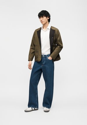 Mladý muž stojí s rukami v vreckách, nosí olivovo zelenú bundu, bielu košeľu, široké modré rifle a biele tenisky na jednofarebnom pozadí.