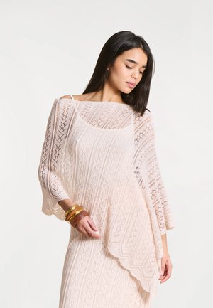 Femme portant un poncho en maille dentelle rose pâle sur une robe assortie, tenant l'ourlet d'une main, et portant des bracelets en or et en bois.